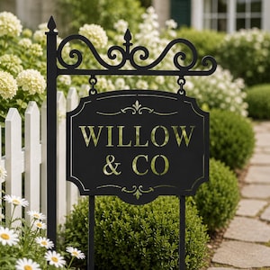 Op de afbeelding: Een hangend bord van zwart metaal met een decoratieve krul aan de bovenkant, gemonteerd op een paal. Het rechthoekige bord bevat de goudkleurige tekst WILLOW & CO in een schreeflettertype, gecentreerd tussen twee kleine gouden decoratieve krullen.