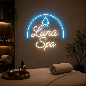 May include: A wall-mounted neon sign displaying the text Luna Spa in white script lettering beneath a blue neon water droplet icon, all enclosed within a blue semi-circular neon arch, illuminating a room with a massage table and spa decor.