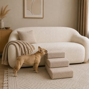 May include: A set of three beige pet stairs with a textured, fuzzy fabric finish. The stairs are arranged in a tiered, step-like structure, positioned on a light-coloured rug in front of a matching beige sofa with a soft, bouclé-style upholstery.