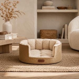 Puede incluir: Una cama redonda para mascotas con un exterior tejido en color tostado y un interior de felpa suave en color crema. La cama cuenta con una base de cojín acolchado y un parche rectangular de cuero marrón con el nombre Daisy grabado en letras oscuras.