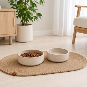 May include: Two round, speckled off-white ceramic bowls placed on an oval, braided tan jute mat. One bowl is filled with brown dry pet food kibble, and the other bowl is filled with clear water. The mat features a small brown leather-like tag.