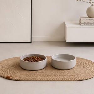 May include: Two light gray cylindrical pet bowls placed on an oval-shaped, tan braided rope mat. One bowl is filled with brown dry kibble, while the other bowl contains clear water. The mat features a small brown leather-like tag on the edge.