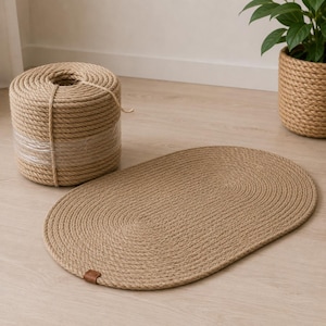 May include: A tan oval-shaped braided rope mat with a small brown leather tag on the edge, accompanied by a large cylindrical spool of matching thick tan rope and a matching rope-wrapped planter containing a green leafy plant.
