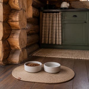 May include: Two round, off-white ceramic bowls placed on a tan, oval-shaped woven rope mat. One bowl is filled with brown dry pet food kibble, while the other bowl contains clear water. The mat features a small brown leather-like brand tag.