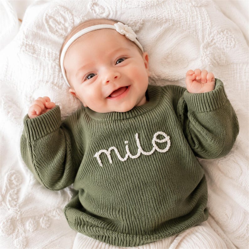 May include: An olive green long-sleeved knit sweater featuring the name "milo" embroidered in white cursive script across the chest. The sweater has a ribbed crew neckline and ribbed cuffs, providing a textured appearance for this garment.