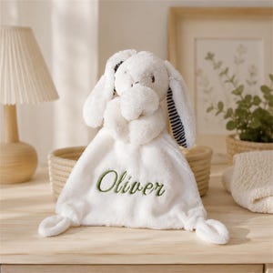 May include: A white plush security blanket featuring a stuffed elephant head with long ears. The ears have a navy and white striped pattern on the underside. The blanket is embroidered with the name Oliver in green cursive script lettering.