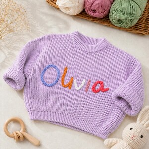May include: A light purple long-sleeve knit sweater featuring the name Olivia embroidered across the chest in multi-colored yarn letters, including blue, orange, white, pink, and red, with a ribbed crew neckline and cuffed sleeves.
