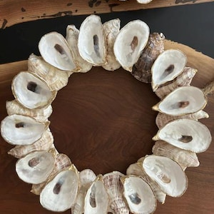 May include: A circular wreath constructed from numerous oyster shells arranged in a ring. Each shell features a smooth, white, pearlescent interior with a dark brown spot, while the exterior surfaces display natural tan and brown textures.