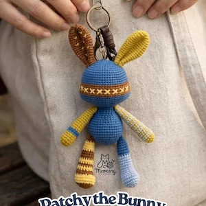 May include: A handmade, crocheted bunny keychain in blue, yellow, and brown. The bunny has long ears, a blue body, and striped arms and legs. The keychain includes a metal clasp and a leather strap. The text "Patchy the Bunny PDF Pattern" is at the bottom.