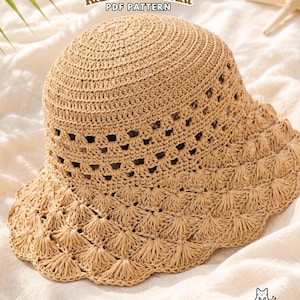 Pode incluir: Um chapéu bucket de ráfia em tom castanho claro com design de crochê, apresentando uma copa circular, uma faixa de pontos abertos e uma aba larga e festonada. O texto "Raffia Bucket Hat PDF PATTERN" está no topo e um logo de gato na base.