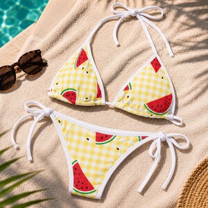 May include: A yellow gingham bikini set with a watermelon print. The bikini top is a triangle style with white ties, and the bottoms are a classic cut with tie sides. Sunglasses are next to the bikini on a beige towel.