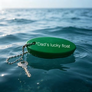May include: An oval-shaped green floating keyring with the text "Dad's lucky float" printed in white. A silver-coloured metal ball chain is attached to the float, connecting to a single standard metal key resting partially in the water.