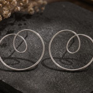 Könnte beinhalten: Ein Paar silberfarbene Ohrstecker mit einem durchgehenden Drahtdesign, das eine große kreisförmige Schlaufe mit einer kleineren, überlappenden Spiralschlaufe in der Mitte bildet. Die Ohrringe haben eine glatte, polierte metallische Oberfläche.