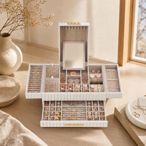 May include: A white multi-tiered jewelry box with a vertical lid featuring an integrated mirror and gold-tone hardware. The interior is lined with light gray fabric and contains multiple compartments for rings, earrings, necklaces, and wristwatches.
