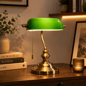 Puede incluir: Una lámpara de banquero con una pantalla de cristal verde vibrante y una base de latón pulido. La lámpara tiene un cuello curvado, un pedestal redondo escalonado y una cadena de metal que cuelga del lateral de la pantalla para encenderla.
