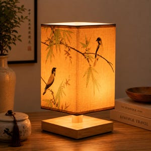 May include: A square table lamp with a light-colored wooden base and a rectangular fabric shade. The shade features a warm, illuminated glow and displays a printed design of green bamboo branches and two small birds perched on the stems.
