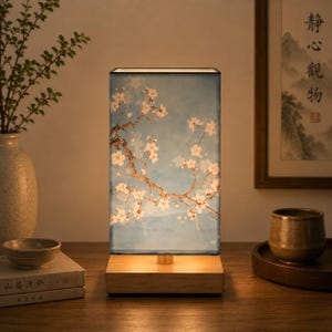 May include: A rectangular table lamp with a light wood base and a shade featuring a blue background with white cherry blossom branches. The lamp emits a warm glow, illuminating the floral pattern printed on the surface of the shade.