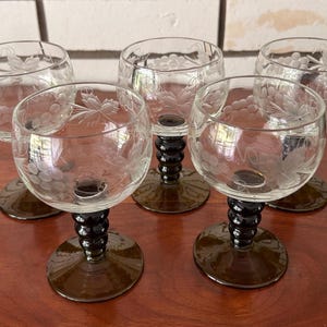 May include: A set of five clear glass goblets featuring etched grape and leaf patterns on the bowls. Each goblet has a dark green, ribbed stem and a circular base. The glasses are arranged in a group, showcasing the transparent bowls and dark stems.