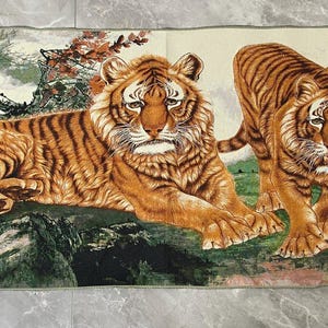 May include: A rectangular tapestry featuring a detailed illustration of two tigers in a landscape. One tiger is lying down on a rocky surface, while the other stands beside it. The scene includes green foliage, dark tree branches, and a mountain backdrop.