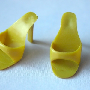 May include: Two yellow plastic doll sandals with a high heel. The sandals have a simple design with a strap across the toes and a small heel. The sandals are a bright yellow color.