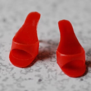 May include: Two small, bright red miniature high-heeled shoes. The shoes have a simple design with a rounded toe and a high, curved heel. The shoes are made of a smooth, glossy material.