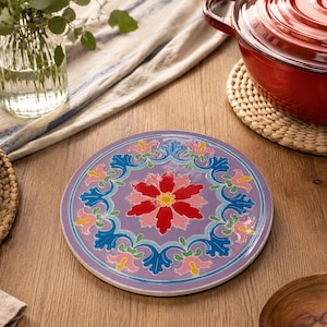 Pode incluir: Um descanso de panela redondo em cerâmica com um padrão floral vibrante, apresentando uma grande flor vermelha central rodeada por um anel de flores estilizadas azuis e cor-de-rosa sobre um fundo violeta claro, concebido para proteger superfícies.