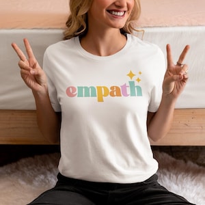 May include: White t-shirt with the word "empath" in pastel colors and star graphics. The shirt is a crew neck style and is made of a soft material. The word "empath" is in pink, yellow, green, and blue.