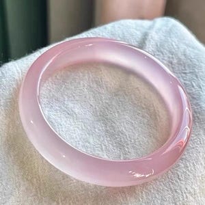 May include: A smooth, translucent light pink bangle bracelet with a polished, rounded surface. The circular jewelry piece features a consistent pale rose hue throughout its solid, continuous band, reflecting light across its curved, glossy exterior.