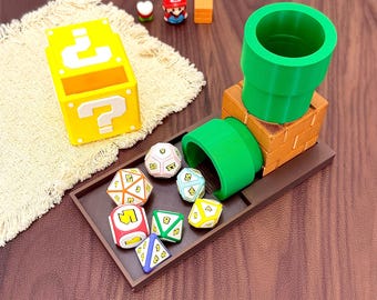 Könnte beinhalten: Ein braunes rechteckiges Tablett mit einer grünen Röhrenstruktur und einem Block mit Ziegelmuster. Ein gelber Fragezeichen-Block steht daneben. Mehrere mehrseitige Spielwürfel in verschiedenen Farben liegen auf dem Tablett.