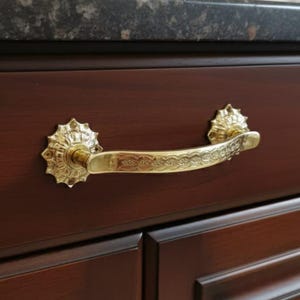 Könnte beinhalten: Ein goldfarbener Schubladengriff mit aufwendigen Details. Der Griff hat einen gebogenen Griff mit einem dekorativen Muster und ist an einem dunkelbraunen Holzschrank befestigt. Die Enden des Griffs haben ein Sonnenstrahl-Design.