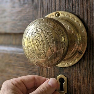Puede incluir: Primer plano de un pomo de puerta antiguo de latón con intrincados diseños florales y de volutas. El pomo redondo está montado en una placa trasera circular a juego, sobre una puerta de madera envejecida. Una mano alcanza la cerradura.