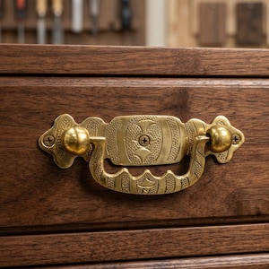 May include: Close-up of a wooden drawer with a decorative brass handle. The drawer is made of dark brown wood with a rectangular frame. The brass handle has a detailed design and two round knobs.