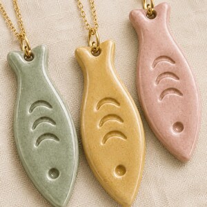 Puede incluir: Tres collares con colgantes en forma de pez en colores verde salvia, amarillo mostaza y rosa empolvado. Cada colgante tiene un acabado liso y brillante con tres hendiduras en forma de media luna y un pequeño orificio circular.