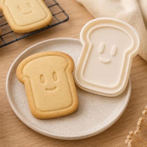 Peut inclure: Un biscuit en forme de tranche de pain avec un visage souriant, à côté d'un emporte-pièce assorti. Le biscuit est sur une assiette, avec d'autres biscuits sur une grille. La palette de couleurs est chaleureuse et accueillante.