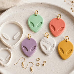 May include: A set of two white plastic alien-shaped clay cutters, five alien-shaped pendants in green, coral, purple, white marble, and yellow, two gold-toned earring hooks, and three small gold-toned jump rings arranged on a round plate.