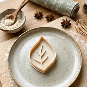 May include: A light beige leaf-shaped cookie cutter with a central vein and branch detail. The cutter is made of a smooth, solid material and features a sharp edge for cutting dough, centered on a speckled, light gray ceramic plate.