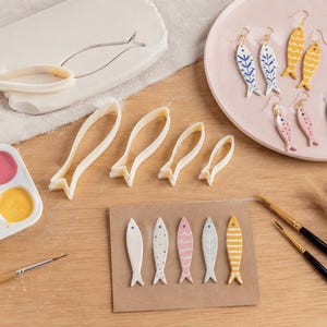 May include: Handmade fish-shaped earrings in various colours and patterns are displayed on a pink plate. Fish-shaped clay cutters and a paint palette with blue, pink, and yellow paint are also visible, suggesting a crafting theme.