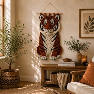 Könnte beinhalten: Ein handgefertigter Tiger-Wandbehang. Der Tiger besteht aus braunem, schwarzem und cremefarbenem Garn. Der Tiger sitzt aufrecht mit seinen Pfoten nach vorne. Der Wandbehang wird an einer Holzstange mit einer cremefarbenen Schnur aufgehängt.