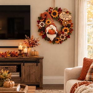 K&ouml;nnte beinhalten: Ein runder Herbstkranz mit geh&auml;kelten Sonnenblumen, roten und orangefarbenen Bl&auml;ttern sowie einer zentralen Wichtelfigur mit hohem orangefarbenem Hut, die einen kleinen orangefarbenen K&uuml;rbis h&auml;lt. Ein Holzschild tr&auml;gt die Aufschrift Autumn Gnome.