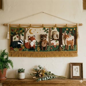 Op de afbeelding: Een rechthoekige gehaakte wandhanger met een bosscène met een das, vos, beer, uil en hert. Het wandkleed hangt aan een houten roede met een wit koord en is voorzien van een mosterdgele rand met franjes aan de onderkant van het kleed.