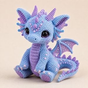 May include: A crocheted dragon figurine in shades of light blue and lavender. The creature features large dark eyes with long lashes, pointed ears, small horns, and wings with subtle sparkling accents. Its body is covered in textured stitches.