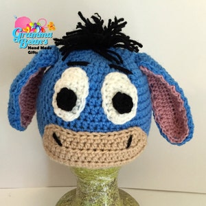 May include: A handmade crochet hat resembling Eeyore from Winnie the Pooh. The hat is mainly blue with white eyes, black pupils, a beige muzzle, and pink inner ears. Black yarn forms Eeyore's hair. The hat is displayed on a mannequin head.