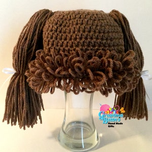 May include: A brown crocheted hat with attached yarn pigtails and a ruffled trim. The hat is displayed on a glass vase. White ribbons secure the pigtails. The text "Grandma Bean's Hand Made Gifts" is visible.