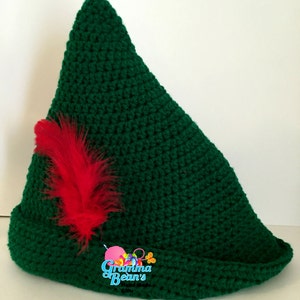 May include: Green crocheted hat with a red feather, shaped like a pointy elf hat. The hat has a brim and is made with a single crochet stitch. The hat is labelled "Gramma Bean's Hand Made Gifts".
