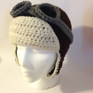 May include: A brown and white crocheted hat with gray and brown goggles attached. The hat has ear flaps.