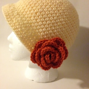 May include: A crocheted hat in a light cream color with a large crocheted flower in a burnt orange color.