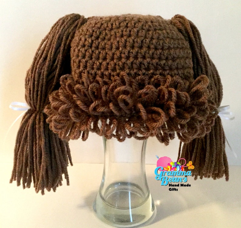 May include: A brown crocheted hat with attached yarn pigtails and a ruffled trim. The hat is displayed on a glass vase. White ribbons secure the pigtails. The text "Grandma Bean's Hand Made Gifts" is visible.