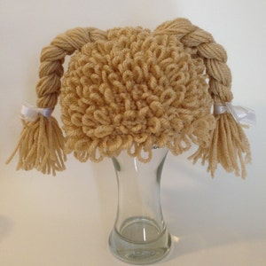 May include: A knitted wig in a light brown colour, featuring two plaited pigtails secured with white ribbons. The wig, made with looped yarn, is displayed on a clear glass vase. It's designed to resemble a child's hairstyle.