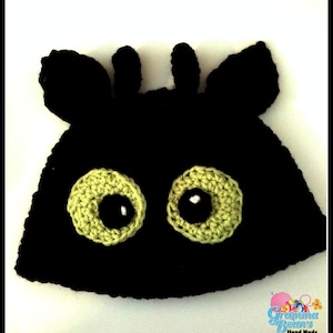 Puede incluir: Un gorro negro tejido a ganchillo con ojos grandes, redondos, de color verde claro y pupilas negras. El gorro tiene dos pequeñas formas erectas en forma de oreja y dos formas más grandes y curvas. El texto "Gramma Bean's Hand Made Gifts" está en la esquina inferior derecha.