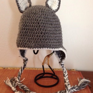 Puede incluir: Sombrero de punto gris con orejas de punto blanco y un largo cordón trenzado blanco y gris.